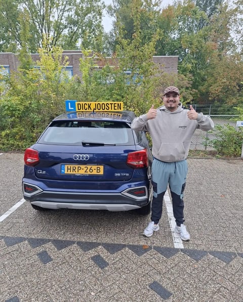 Parham steekt zijn duimen op naast de blauwe Audi-lesauto na zijn examen