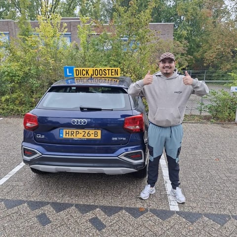 Parham steekt zijn duimen op naast de blauwe Audi-lesauto na zijn examen